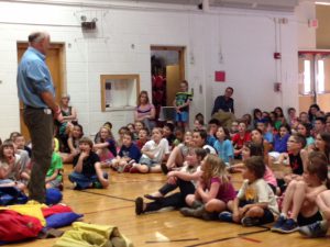 Author in the gym talking to students who are sitting on the gym floor.