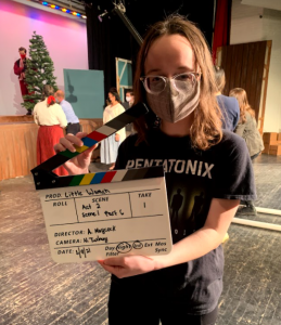 masked student standing on a stage holding a clapperboard for filming for Little Women. Costumed cast in the background.