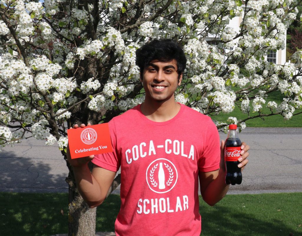 Guilderland High School Senior Aneesh Muppidi, standing in front of a flowering tree with white flowers and wearing a red t-shirt with white lettering that reads "Coca-Cola Scholar", smiles for the camera and holds a card with the words "celebrating you" and a bottle of Coca-Cola soda.