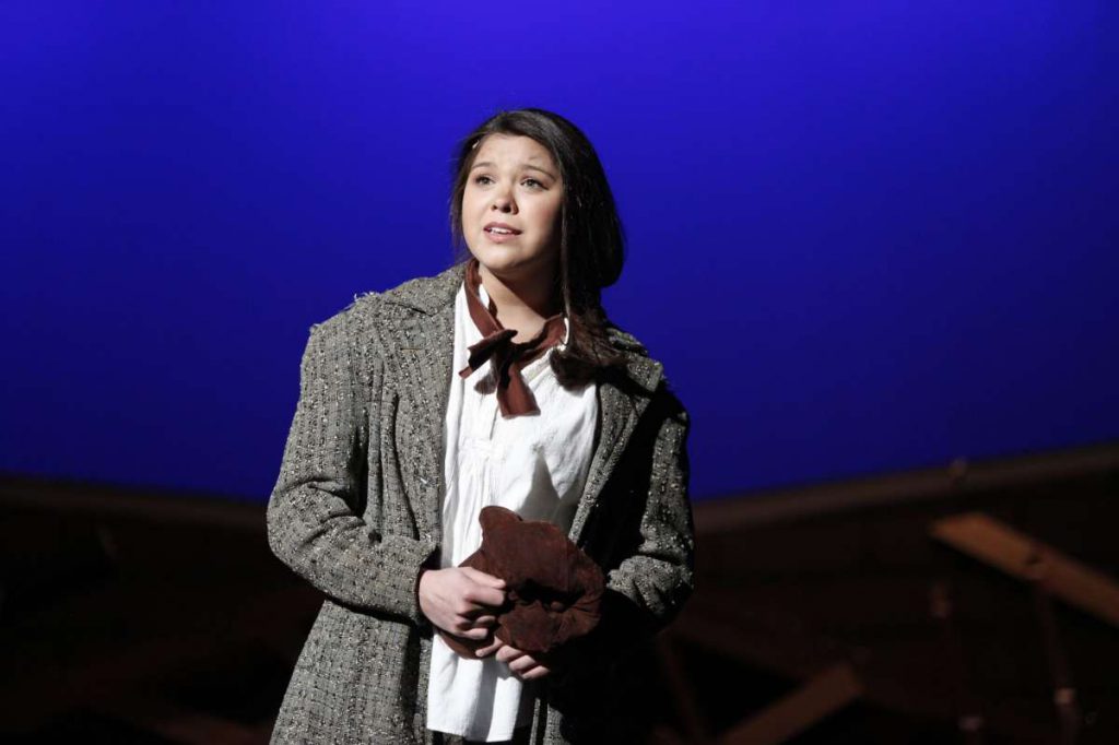 Guilderland High School student Nicaya-Isabella Rios, dressed in white blouse, brown scarf and oversized tweed jacket, performs onstage. The stage is lit in a bright blue.