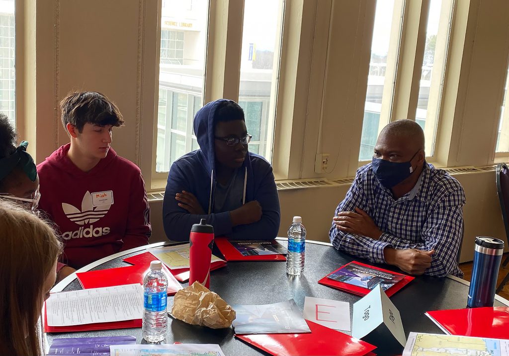 2 male high school students are seated next to an a male native speaker from Togo. There's a sign on the table that says "Togo" and each student has red folders in front of them, as well as assorted drinks.