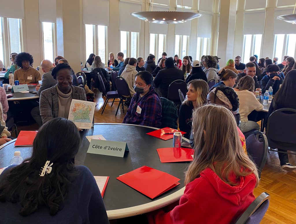 6 female GHS students sit a table that says "Ivory Coast" in French and are discussing the country's culture. Several other tables are filled with students in the background doing the same thing. 