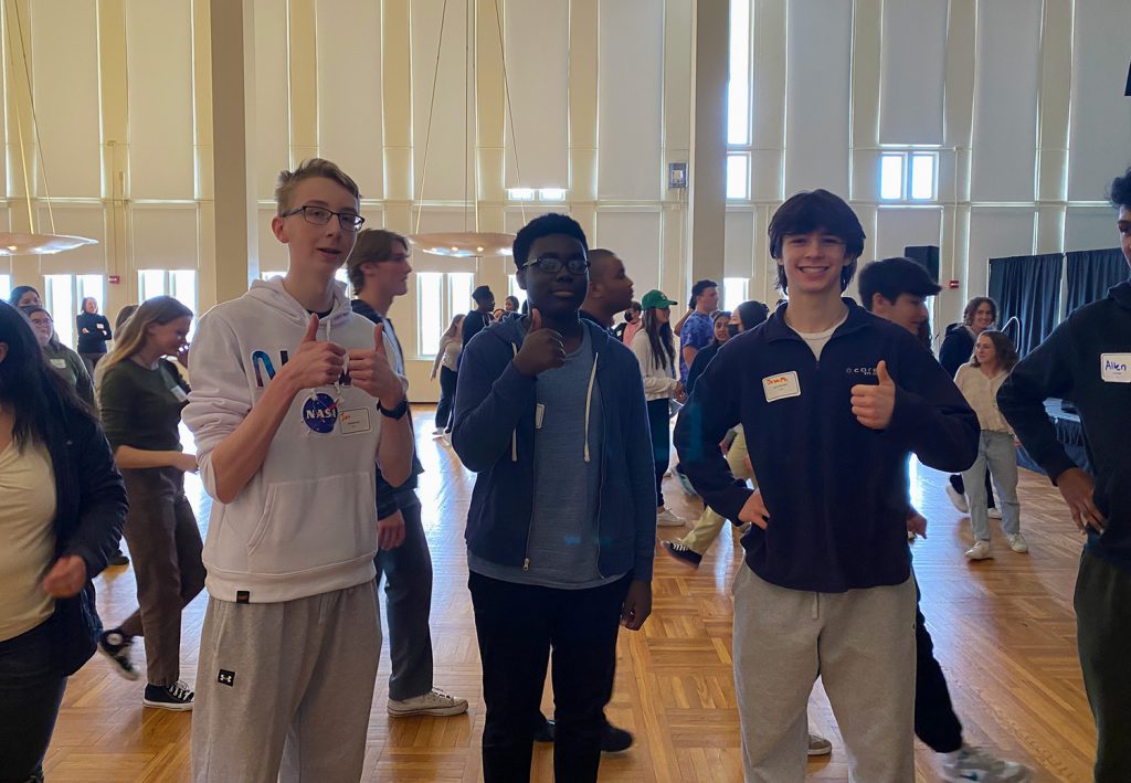 3 male GHS students are standing next to each other and each is giving a "thumbs up" as they smile at the camera. The background consists of several other high school students in attendance at the event.