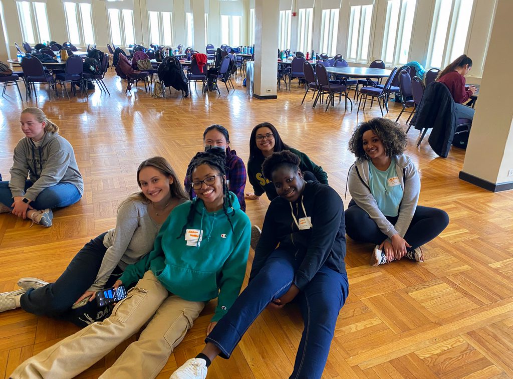 6 female GHS students are sitting on the hardwood floor smiling at the camera for a picture. The background consists of several tables with chairs, with no one sitting in them, but the chairs have people's belongings on them.