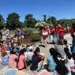 Student volunteer wearing red t-shirt holds up a book to children seated in a semi-circle around her.