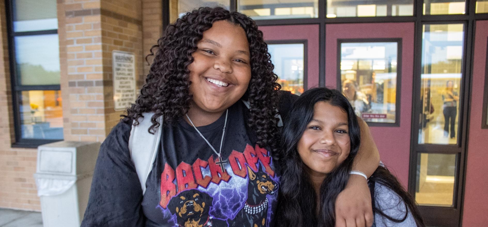 Two students with their arms around each other in front of high school entrance, smiling for a picture on the first day of school
