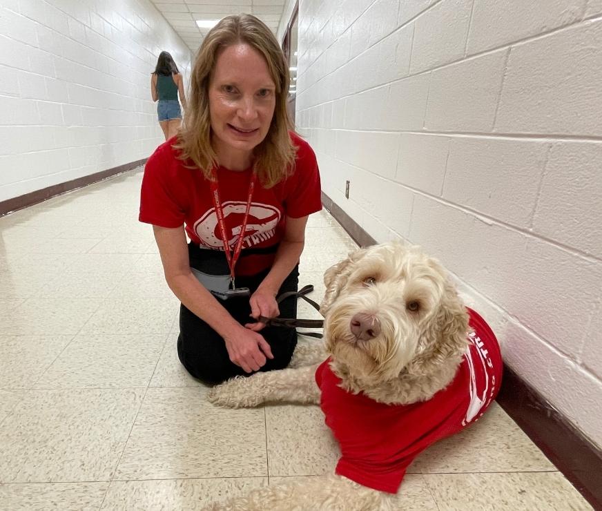 Staff member in red Guilderland shirt poses with white haired therapy dog also in red Guilderland shirt.