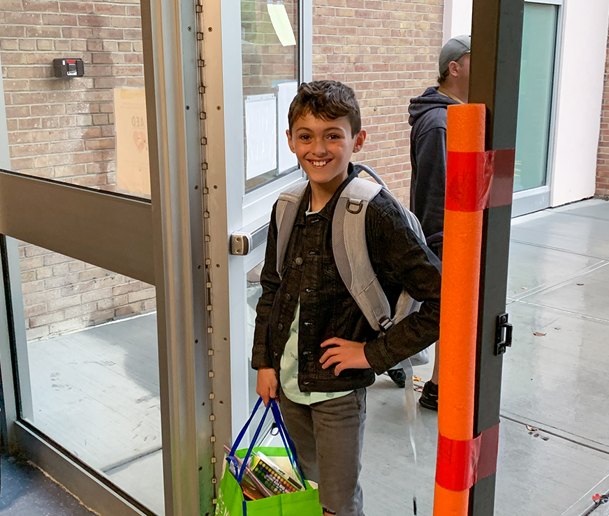 An elementary student walking into school wearing a backpack, smiling.