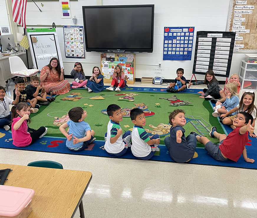 Students and teacher sitting on the floor, smiling and giving thumbs up.