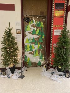 A door decorated with paper trees and a snowy path made from paper. The door is flanked by Christmas trees, on either side.