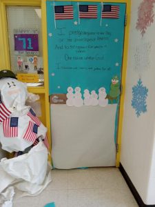 A door decorated with flags and snowmen. The pledge of allegiance is written on the paper covering the door. Next to the door is a snowman made of paper. The snowman has flags on it.