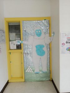 A door decorated with a snow monster, made from white and light blue fabric. The snow monster has snowballs at this feet and one in each hand.