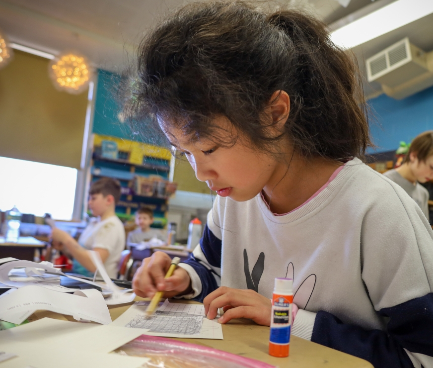 Student sits at desks and works on math tables with a pencil.