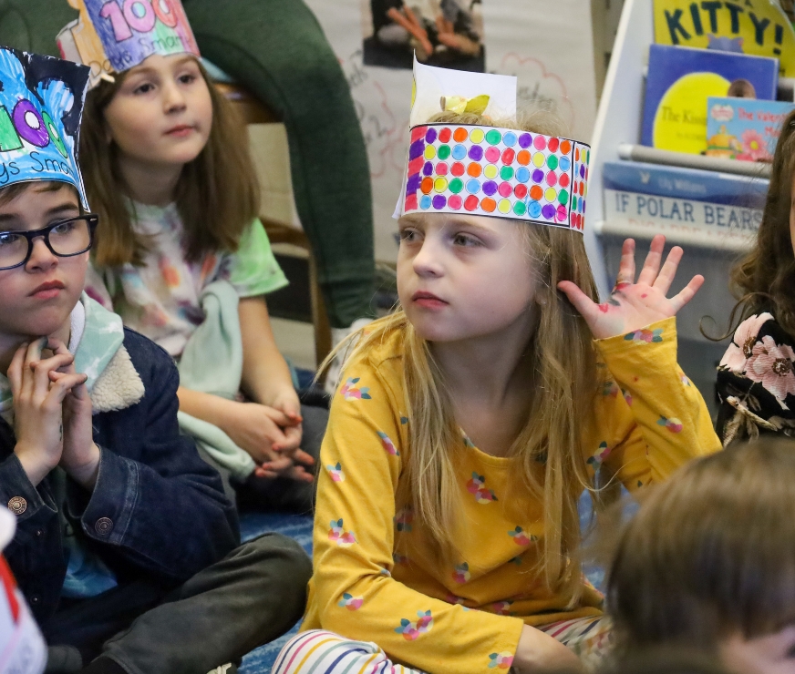 Student sits on the ground raising hand, while wearing decorative hat for 100th day of school.