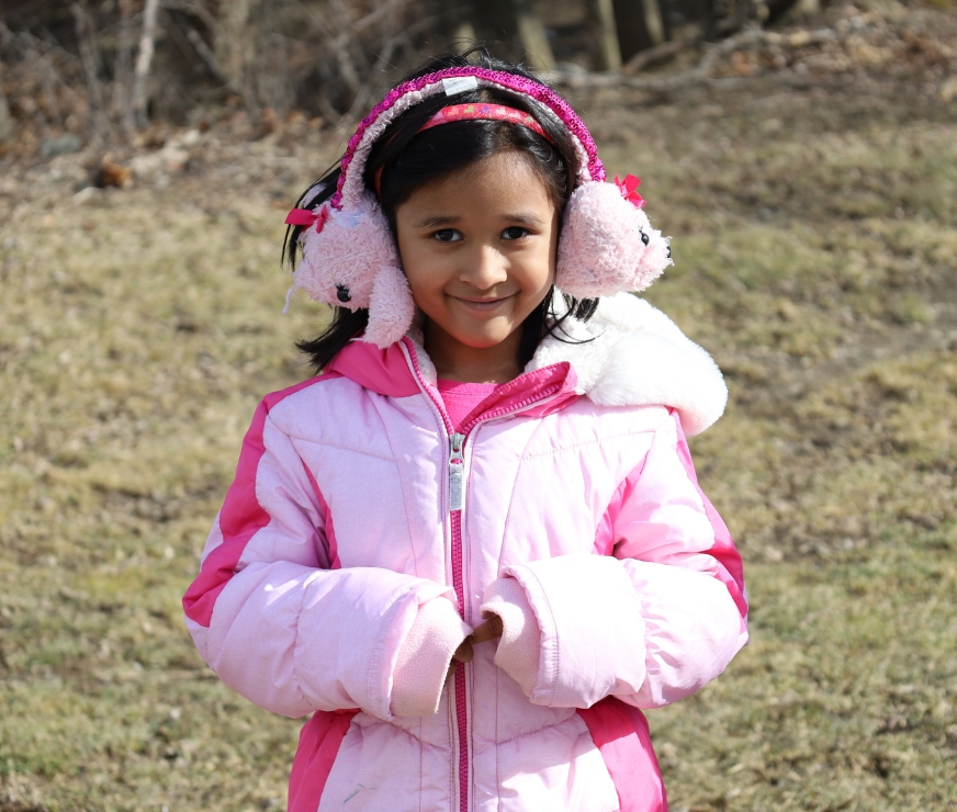 Student outside wearing coat and earmuffs looks at the camera and smiles.