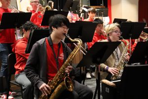 Two students play saxophone at the Young People's Concert. One is wearing a red t-shirt, one is wearing a tan colored jacket. Other band members are behind them, two are playing the trumpet.