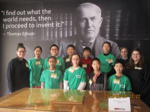 A group of students, some wearing bright green t-shirts, and two adult advisors, stand in front of a black and white image of Thomas Edison. Nex to the image is a quote that reads: I find out what the world needs, then I proceed to invent it.