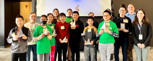 Group of about 10 students and a few adults. The students are holding triangular shaped awards, several are wearing bright green t-shirts.