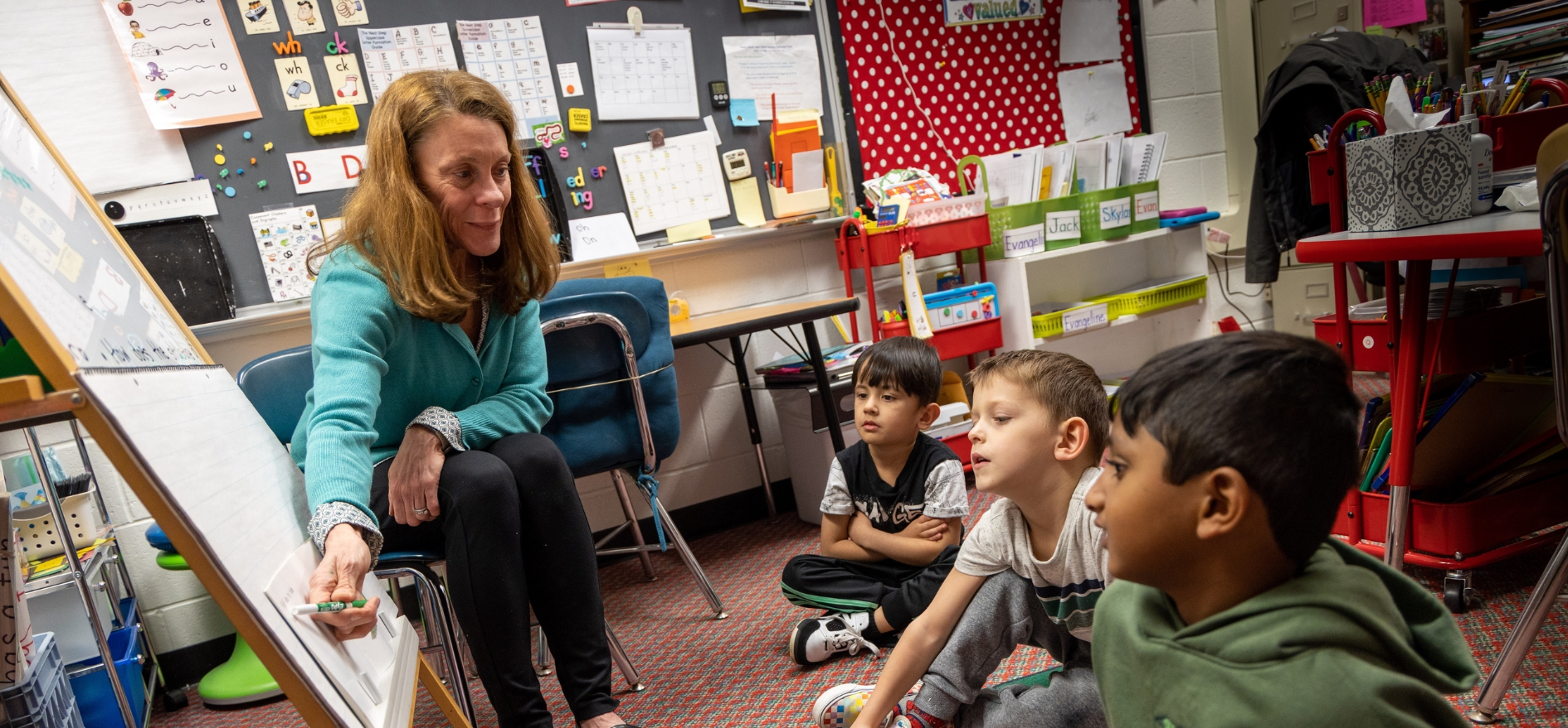 Teacher sitting in chair points to board, walking students through how to put together a word.
