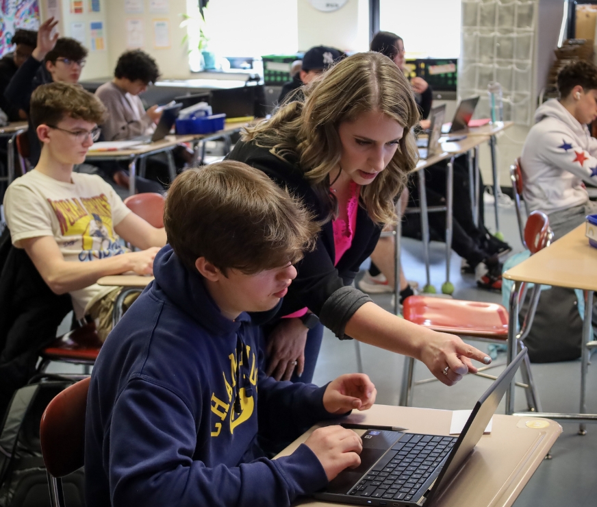 A teacher points out a detail to a student on a laptop.