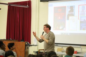 Visiting author gestures with his right hand. He is holding a microphone with his left hand. He is standing in front of a white screen, projected on the screen are the images of book covers to the books he has written.