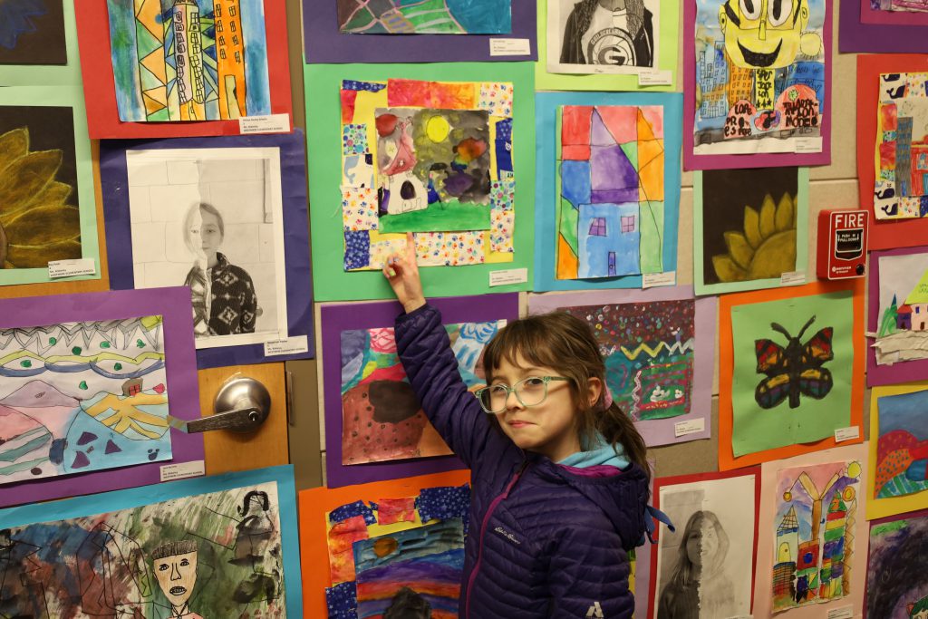 Elementary student points to a piece of artwork she created, in front of a wall of colorful artwork.