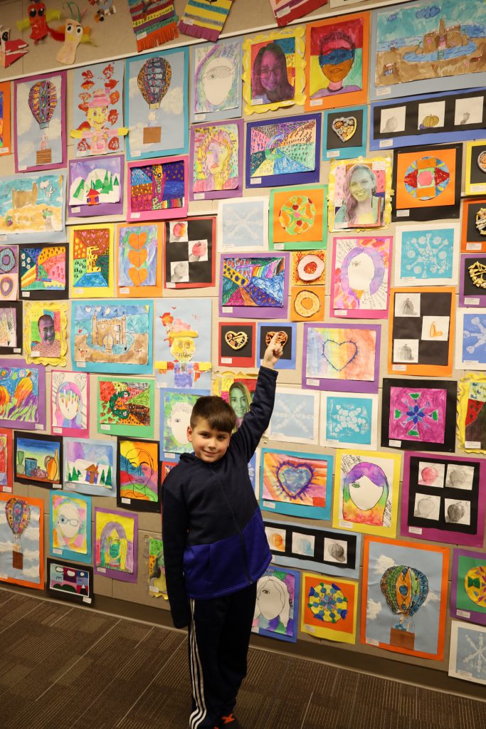 Student stands in front of a wall of colorful artwork and points to a piece he created.