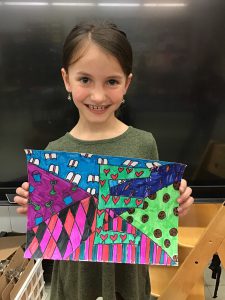 Elementary students stands in front of blackboard, holding up colorful artwork