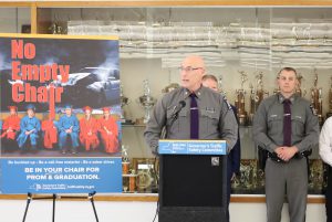 NYS Police Major speaks at a podium in front of a line of state police officers. He stands in front of a trophy case and next to a poster that reads no empty chair.