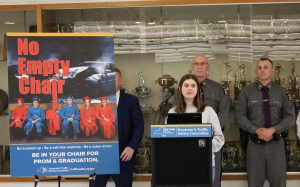 Student speaks at a podium in front of a line of state police officers. She stands in front of a trophy case and next to a poster that reads no empty chair.