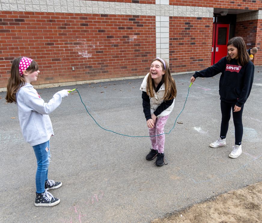 Student laughs as they jump rope with two friends holding each side of the rope.