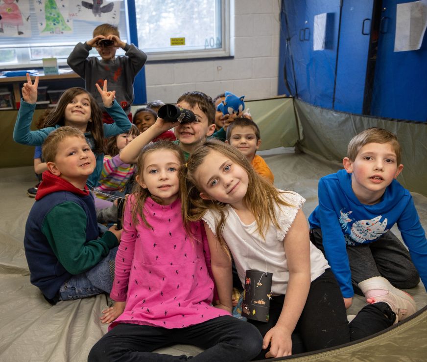 Students pose together in classroom camping tent. Student in the middles looks through binoculars.