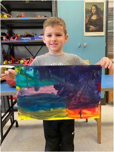 Elementary student stands in classroom, holding colorful artwork