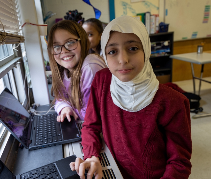 Students stand by window and work on their laptops.