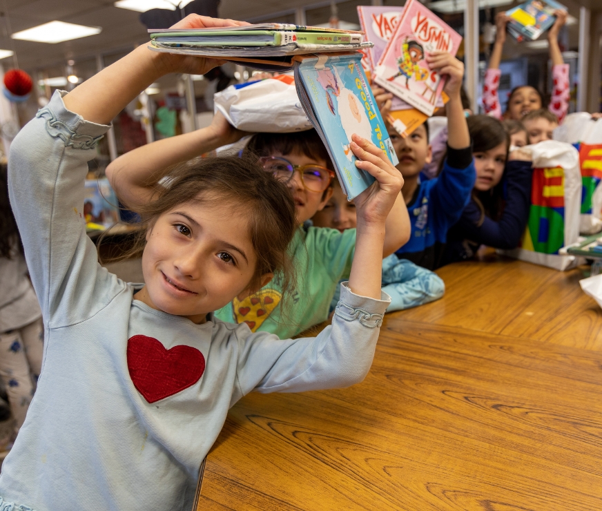 Students pose for a picture by holding books over their head as they stand next to library counter.