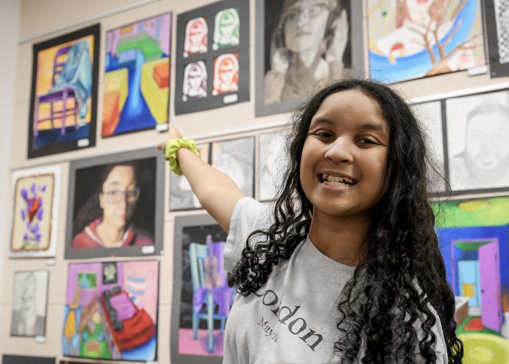 Student points to artwork on a wall where many pieces of artwork are displayed.