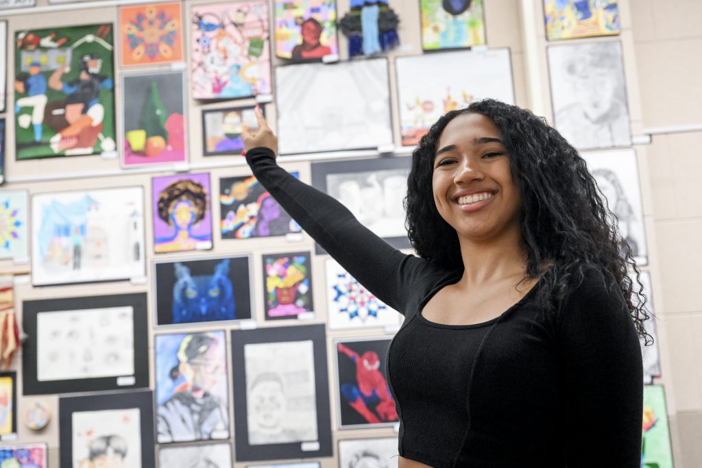 Student points to artwork on a wall where many pieces of artwork are displayed.