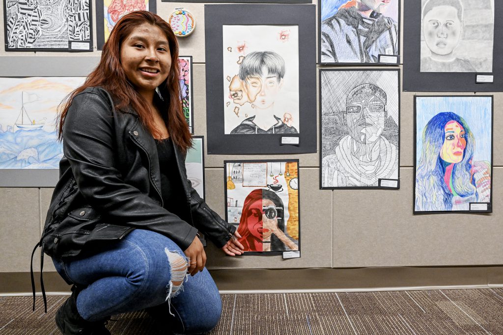 Student points to artwork on a wall where many pieces of artwork are displayed.