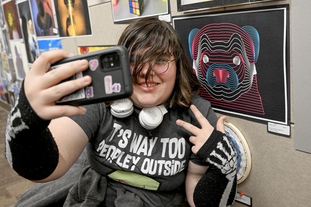 Student holds a cell phone in front of them to take a selfie with artwork.