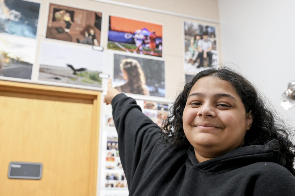 Student points to artwork on a wall where many pieces of artwork are displayed.