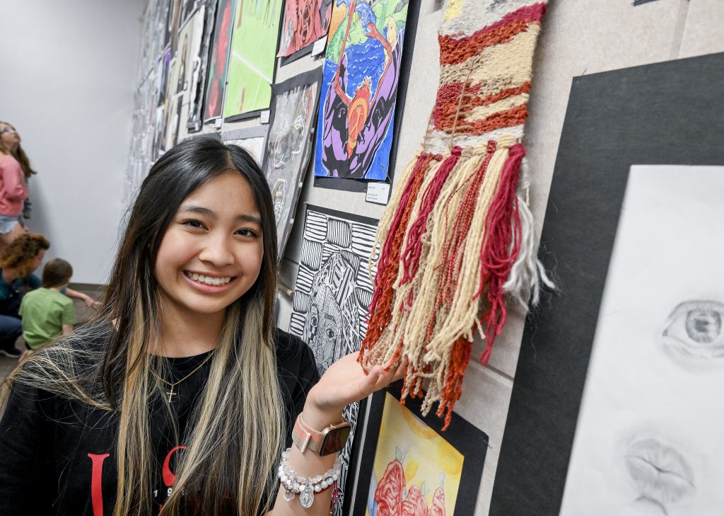 Student smiles while posing next to three-dimensional piece of art hanging on the wall.