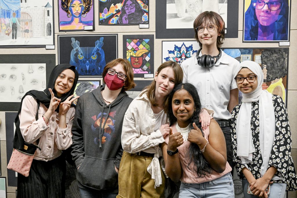 A group of students pose in front of artwork. Some are giving thumbs up, some are giving peace sign.
