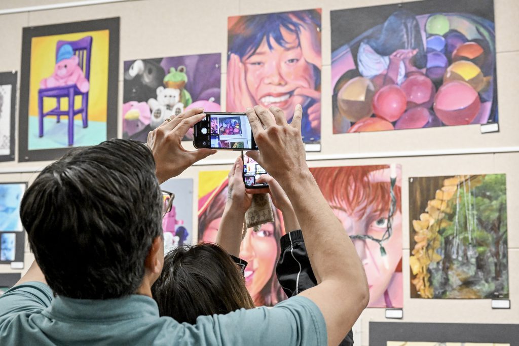 People are taking photos of artwork on display. You can see artwork on the cell phone screen.