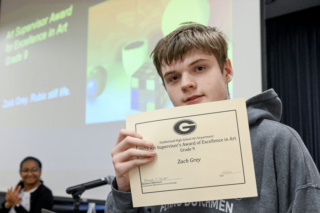 Student holds up certificate in a close-up photo. Behind the student is a screen displaying artwork.