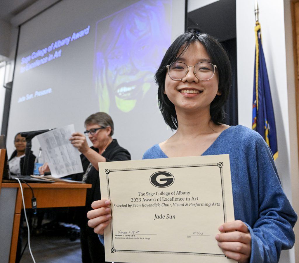 Student is smiling, holding certificate. Behind the student is a screen displaying artwork and person at a podium.