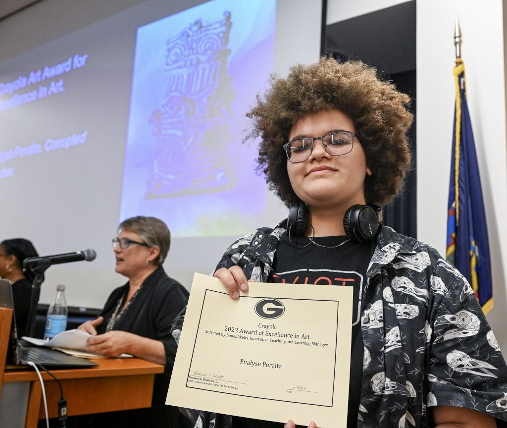 Student is smiling, holding certificate. Behind the student is a screen displaying artwork and person at a podium.