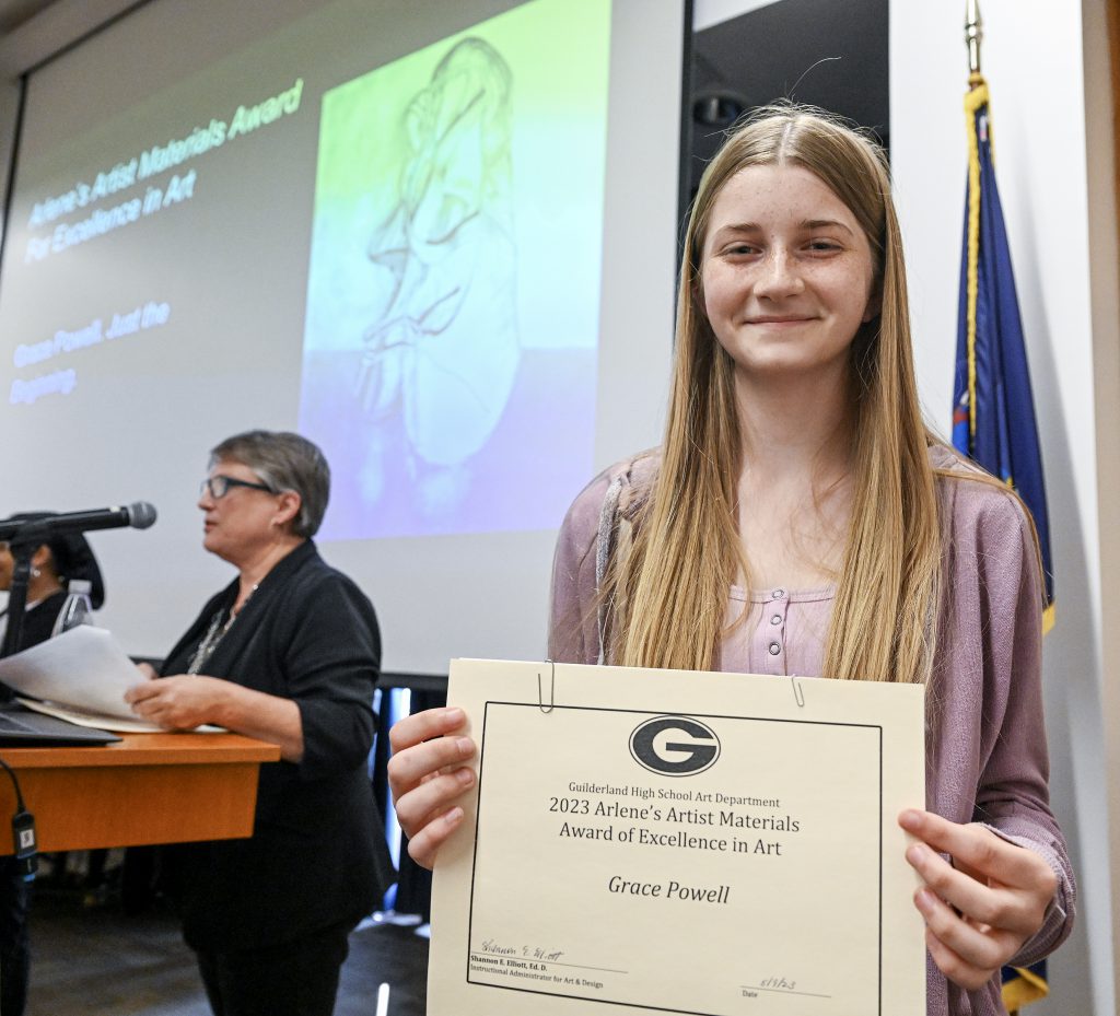 Student is smiling, holding certificate. Behind the student is a screen displaying artwork and person at a podium.