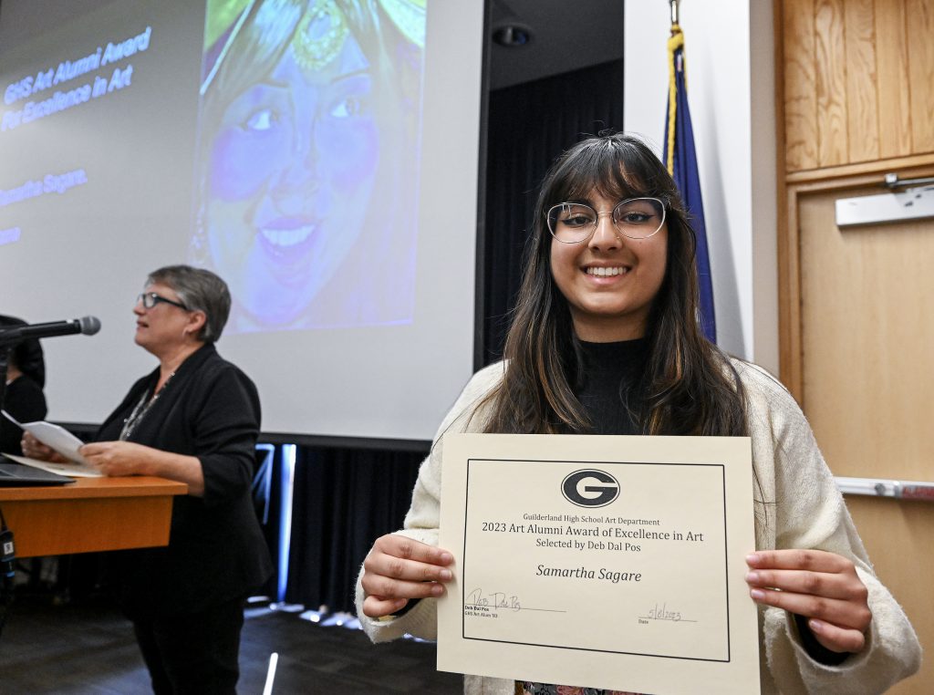 Student is smiling, holding certificate. Behind the student is a screen displaying artwork and person at a podium.