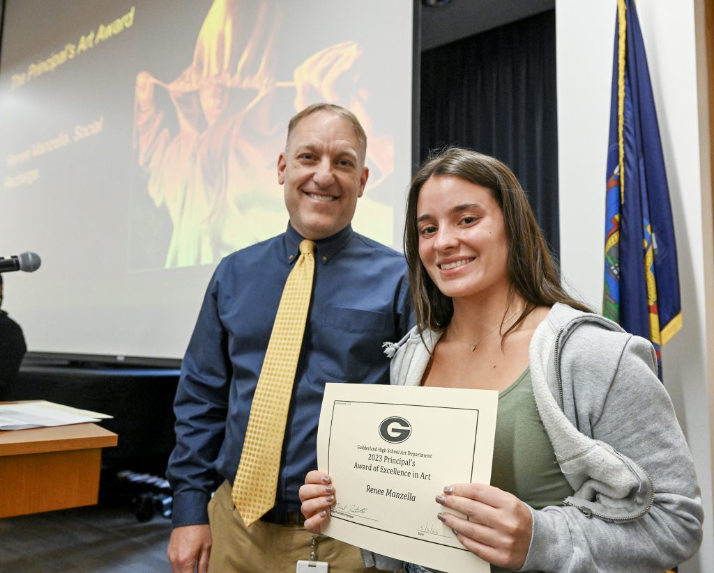 Student is smiling, holding certificate and posing with GHS principal. Behind the student is a screen displaying artwork and person at a podium.