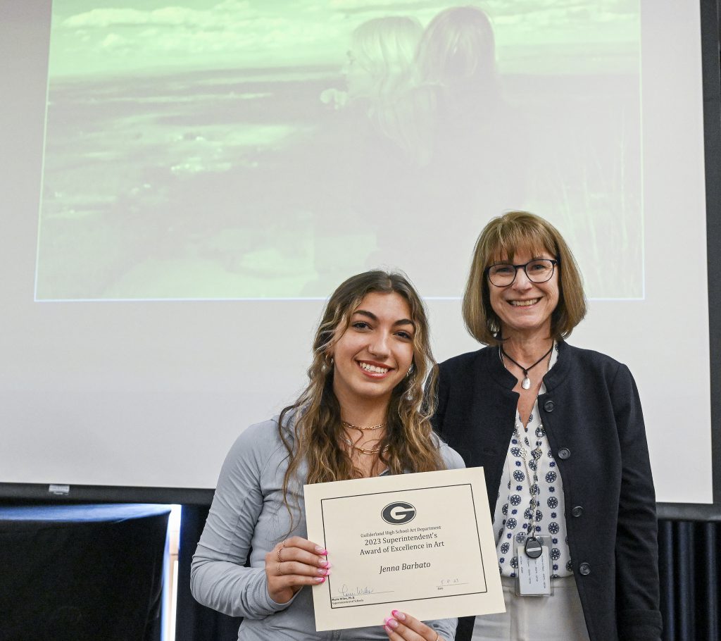 Student is smiling, holding certificate, posing with GCSD superintendent. Behind the student is a screen displaying artwork and person at a podium.